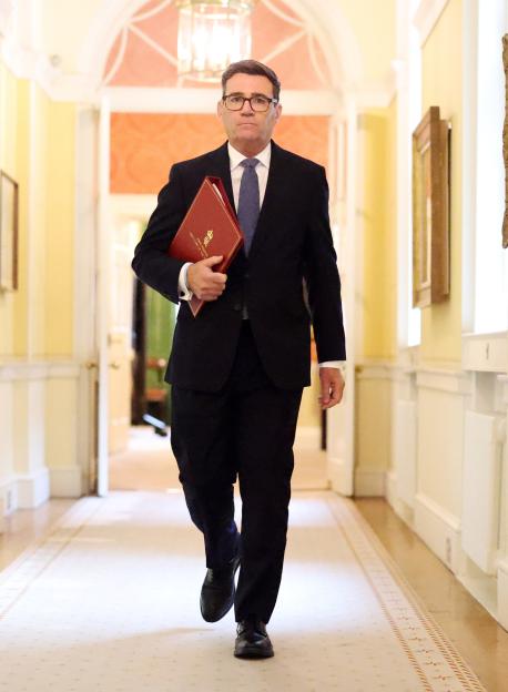 Prime Minister Andy Burnham walks down a hallway at 10 Downing Street holding a red folder.