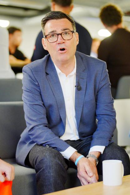 Andy Burnham, in a blue suit jacket and white shirt, sits with an open mouth, wearing glasses and a microphone clipped to his shirt.