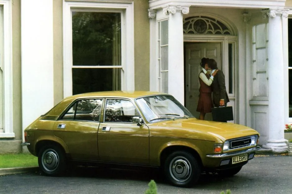 A mustard-colored Austin Allegro parked in the driveway of a grand house, with a couple kissing in the doorway.