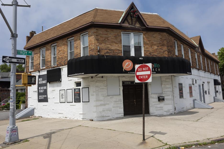 A brown brick building with a white first floor, a black awning, and a "Do Not Enter" sign in front of its main entrance.