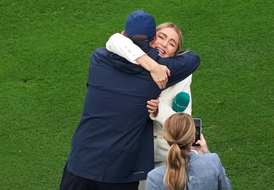 A woman reporter with a microphone hugs a man, while another woman takes a photo of them.