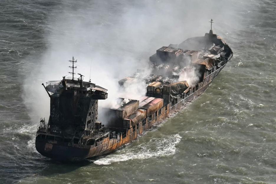 A cargo ship burns, billowing smoke from its deck, in choppy waters.