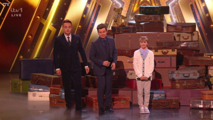 Two male hosts and a young boy stand on stage in front of a backdrop of stacked vintage suitcases.