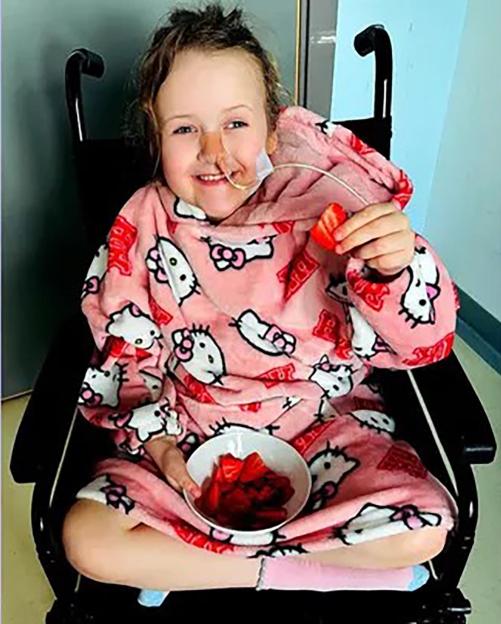 A smiling young girl in a pink Hello Kitty onesie, with a feeding tube on her face, holds a strawberry while sitting in a wheelchair.