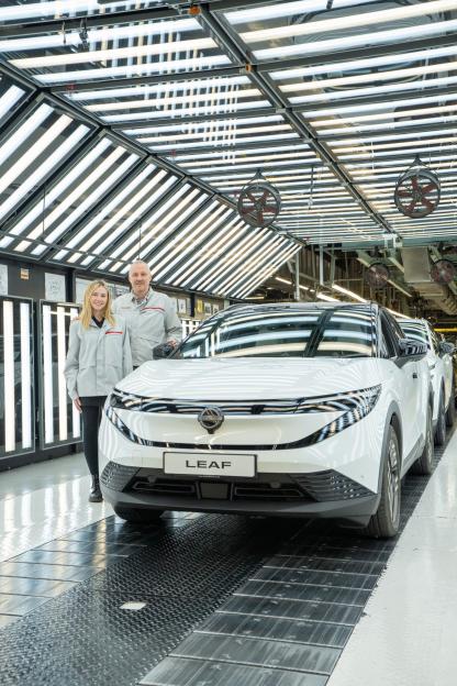 Two people in factory uniforms stand beside a white Nissan Leaf electric car in a brightly lit factory.