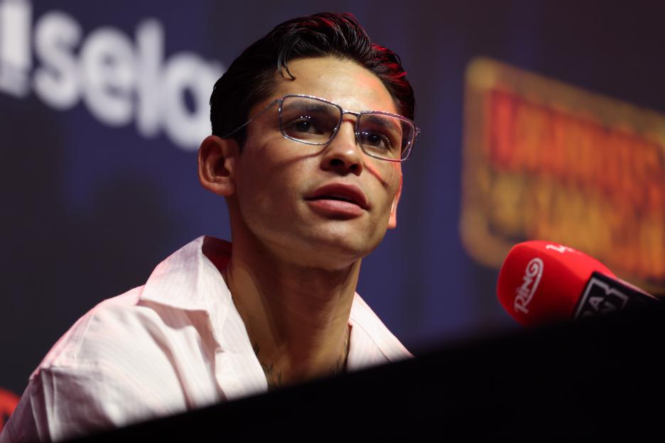 Ryan Garcia speaks at a press conference, wearing glasses and a light-colored shirt, with a microphone in front of him.