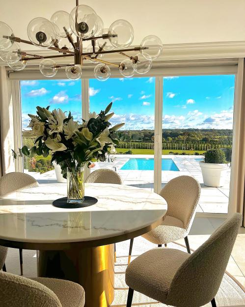 A dining room with a white marble table and a large bouquet of white lilies, overlooking a pool and green landscape under a blue sky.