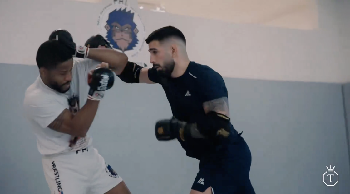 Two men sparring in a gym, one wearing a black shirt and the other a white shirt.
