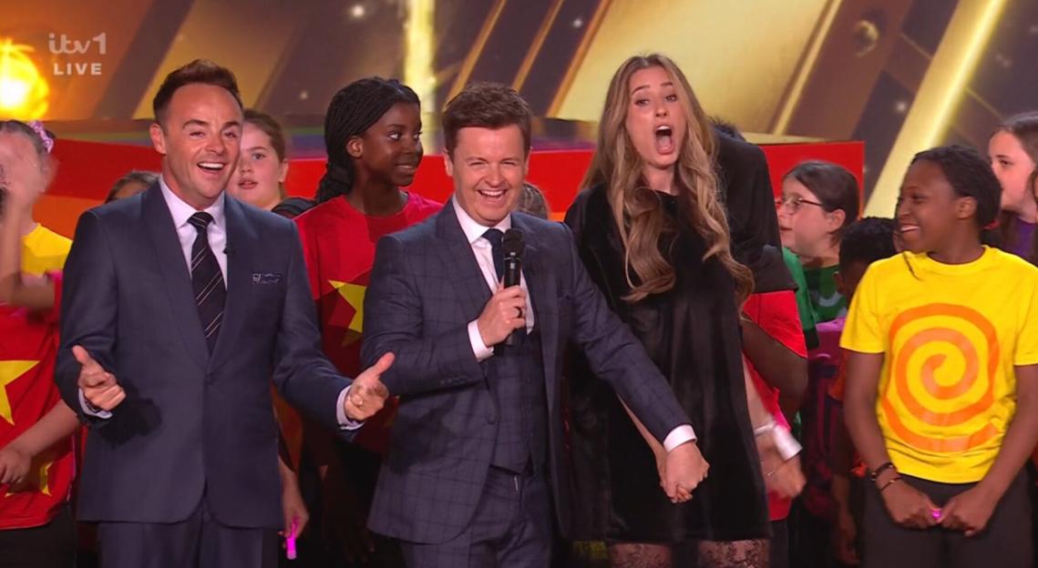 Two hosts on stage at Britain's Got Talent, one with a microphone, surrounded by children and a woman.