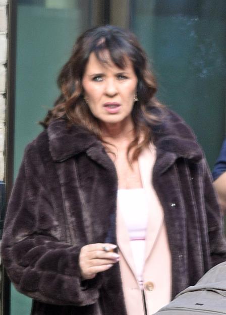 Coleen Nolan smoking a cigarette outdoors, wearing a dark fur coat over a light pink top.