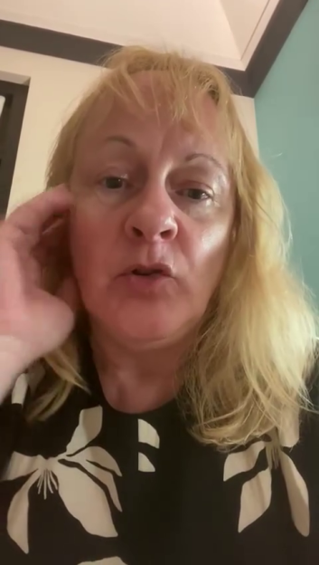 A blonde woman in a black and white floral top looks directly at the camera.