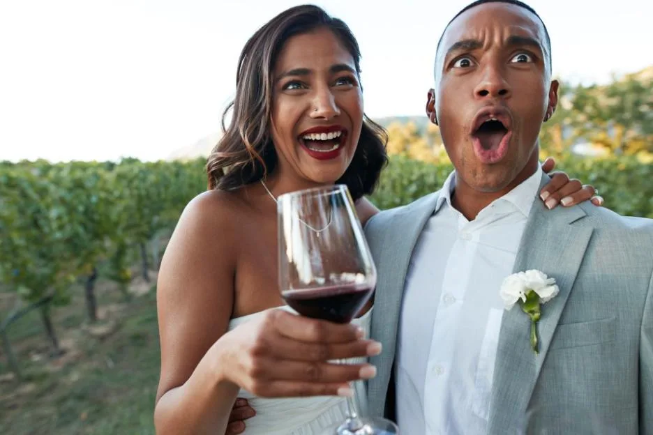 A shocked groom stands next to a happy bride holding a wineglass in a vineyard at sunset.