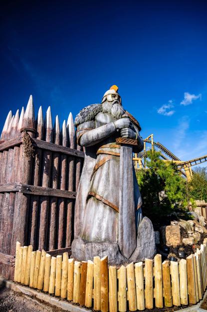 A large statue of a Viking warrior holding a sword, next to a wooden fence, with a roller coaster in the background.