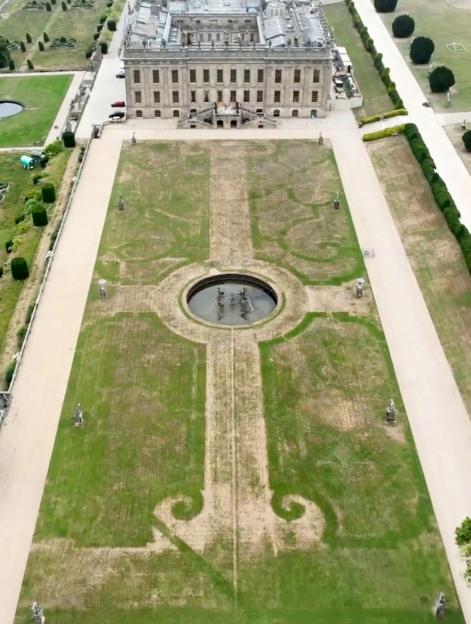 Aerial view of a stately home with a large lawn in the foreground showing parched lines revealing a historic garden design underneath.