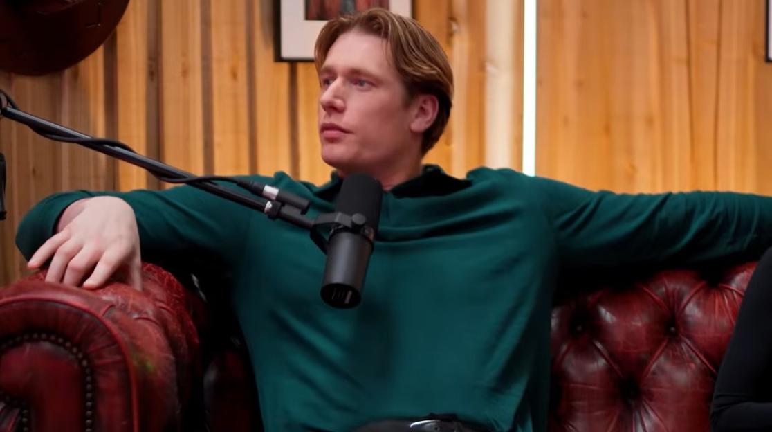 A man in a green shirt sits on a red leather couch with a microphone in front of him.