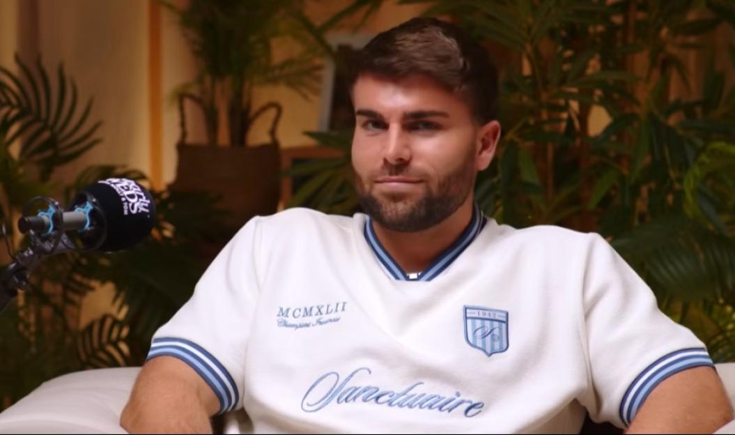 A young man in a white sweater with blue stripes and the word "Sanctuaire" embroidered on it, sitting in a podcast setup with a microphone in front of him.