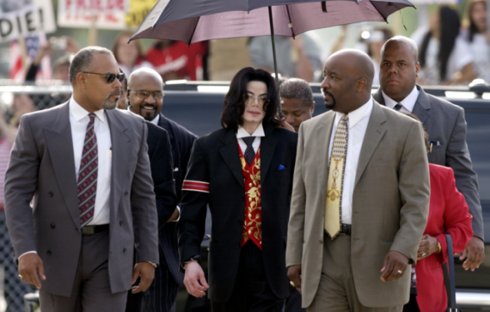 Michael Jackson with long dark hair, wearing a black suit, red vest with gold embroidery, and glasses, walks surrounded by his entourage, with one person holding an umbrella over him.