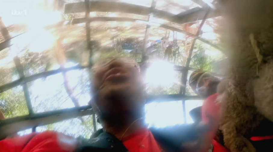 Two people, seen from below, drenched by water, with one looking up and the other wearing a cap, surrounded by wooden beams and bright light.