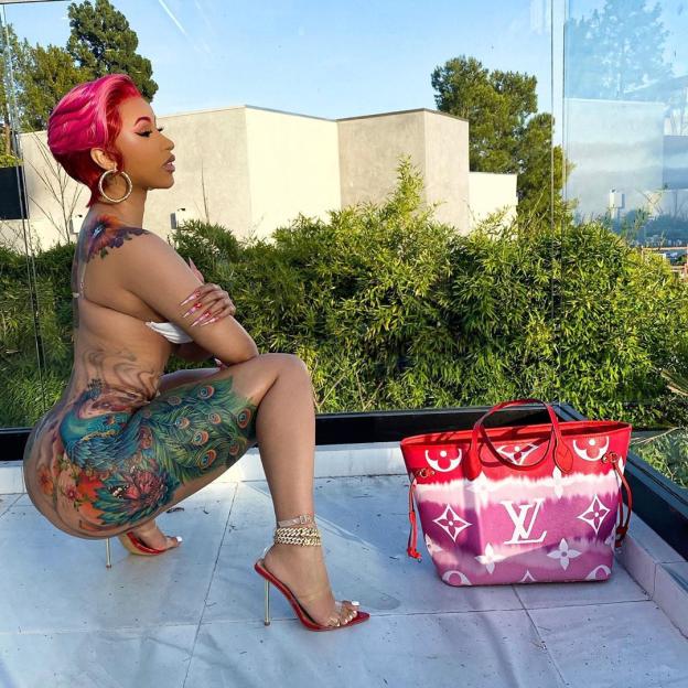 Cardi B with a large, colorful peacock tattoo on her leg, next to a red and pink Louis Vuitton bag.