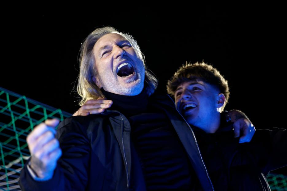 A man with long blonde hair and a younger man with curly brown hair joyfully celebrating under blue lighting at a football match.