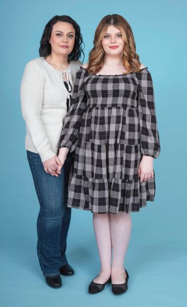 A woman in a white sweater and jeans holds hands with a younger woman in a plaid dress, both looking at the camera.
