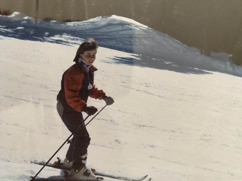 A woman in a red and blue ski jacket and dark pants skiing down a snowy slope, holding ski poles.