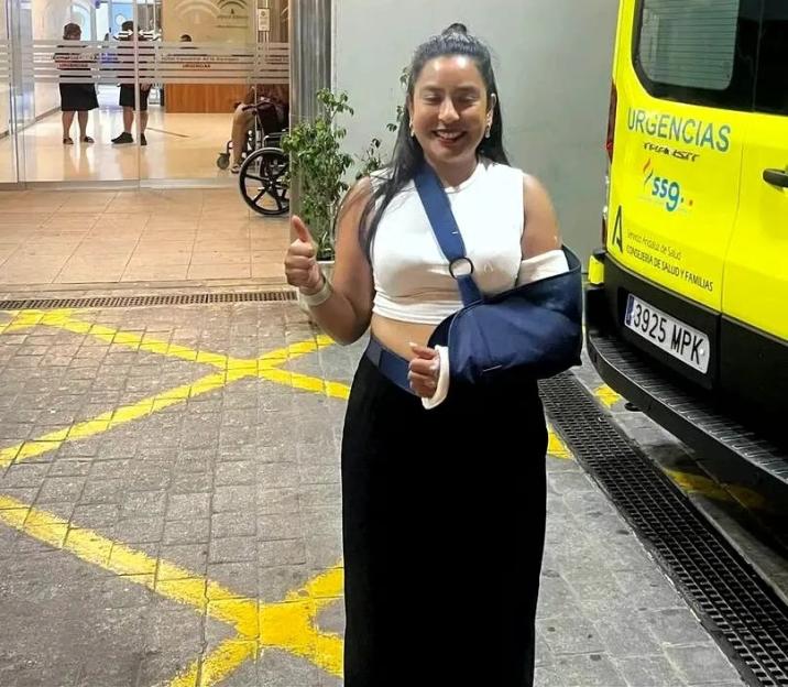 A woman with her arm in a sling giving a thumbs up in front of a hospital entrance and an ambulance.