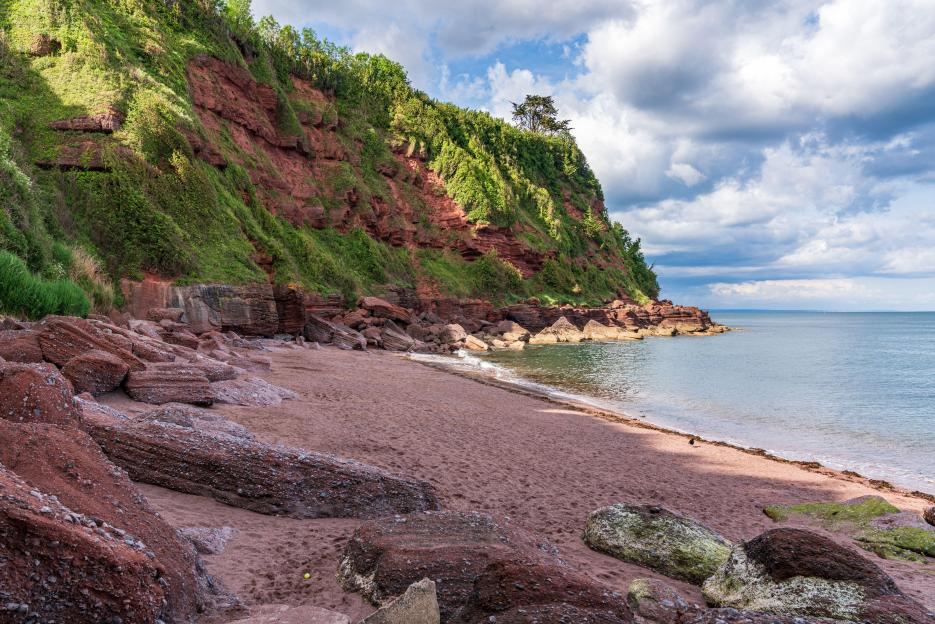 Maidencombe beach in Torbay, England, showing red cliffs, sandy shore, and the sea under a cloudy sky.