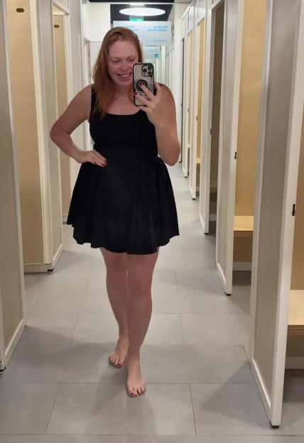 A woman in a black mini-dress stands barefoot in a dressing room hallway.