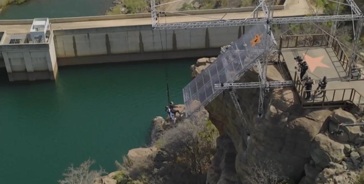 A person in a harness suspended by a rope above a lake, next to a large metal structure with a star logo.