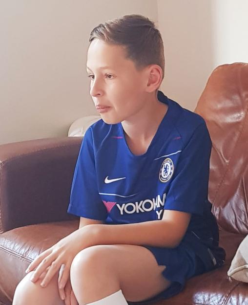 A boy, Louis Watkiss, wearing a blue Chelsea football shirt, sits on a brown leather sofa.