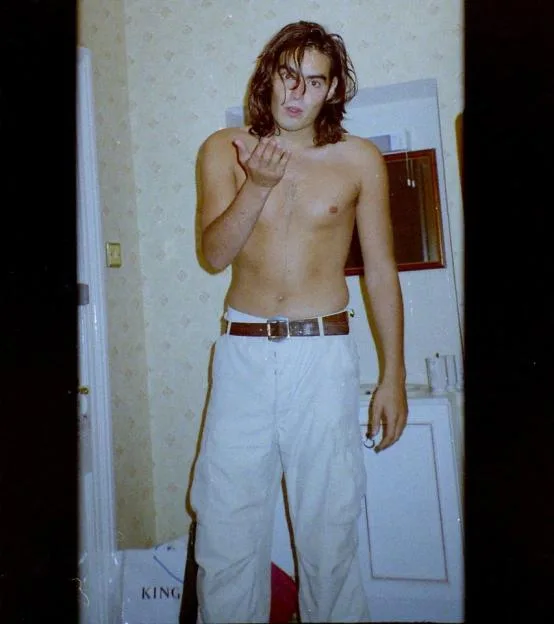 A shirtless British comedian Russell Brand with long hair, wearing white trousers and a belt, blowing a kiss in a hotel room with a Metropolitan Police logo in the top left corner.