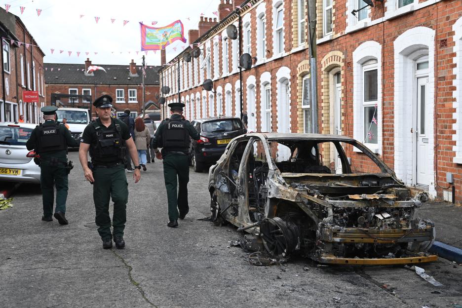 Police walk past a burnt-out car on Lendrick Street in Belfast following anti-immigrant riots.
