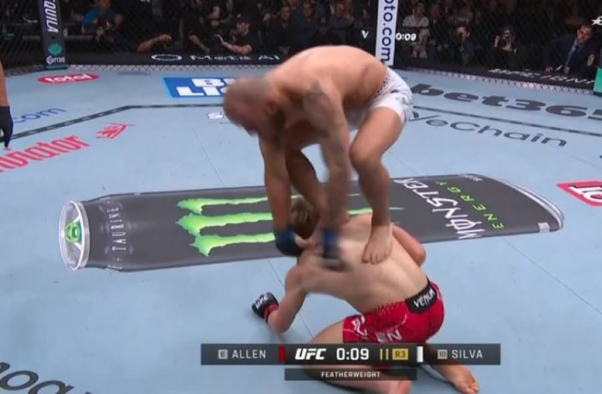 A fighter in white shorts stomps on an opponent in red shorts, who is on hands and knees, in a UFC octagon.
