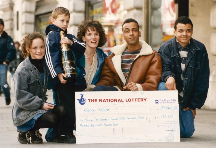 National Lottery winners Karren Taylor and Lee Ryan with their three children, holding a large check for £6.5 million and a bottle of champagne.