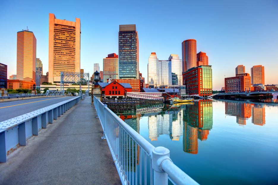 The Boston skyline at sunset with buildings reflecting in the harbor.
