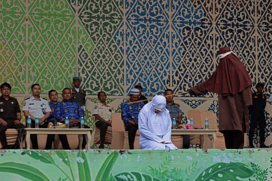 Banda Aceh, Aceh, Indonesia. 17th Sep, 2026. A hooded Acehnese woman kneels to receive lashes from a masked sharia officer as officials and law enforcement personnel watch in the background during a public caning execution at Taman Sari in Banda Aceh