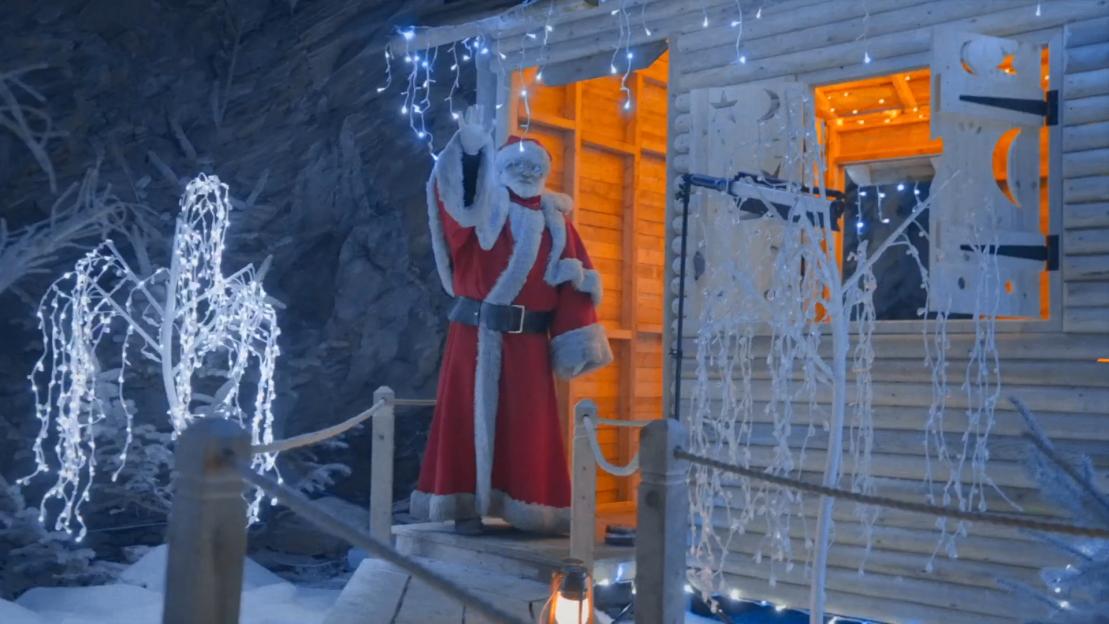 Santa Claus waves from a wooden house decorated with Christmas lights.