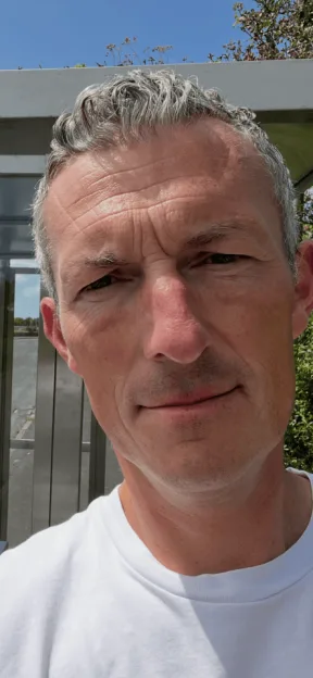 A close-up selfie of a middle-aged man with graying hair and a neutral expression outdoors in bright sunlight.