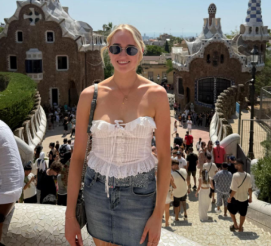 A woman in sunglasses and a white top smiles in front of Antoni Gaudí's gingerbread houses in Park Güell, Barcelona.