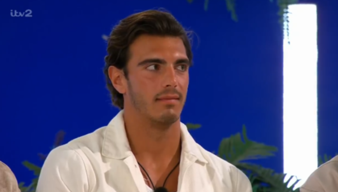 A young man with dark hair and a beard, wearing a white shirt, looks to the right against a blue background.