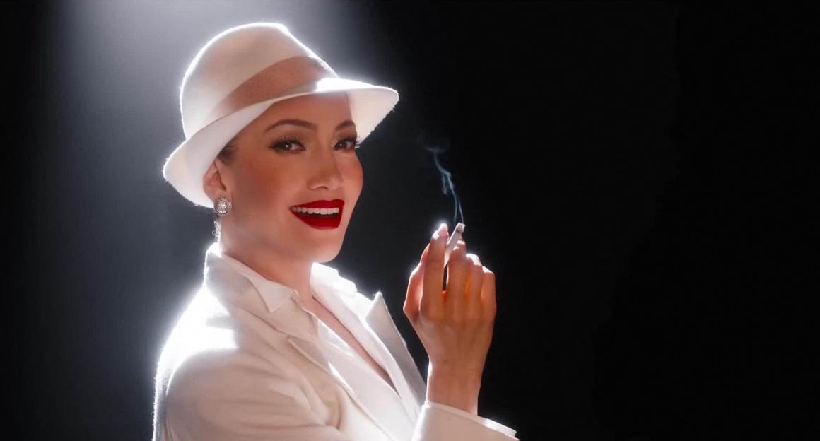 Jennifer Lopez as Aurora in "Kiss of the Spider Woman" with red lipstick and a white hat, holding a smoking cigarette.