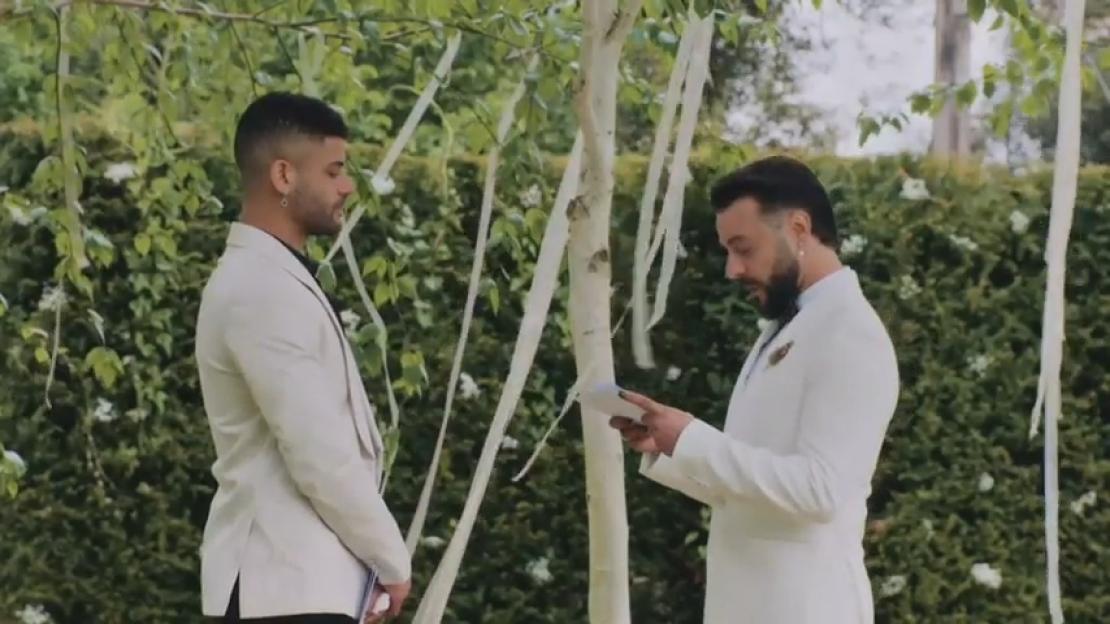 Groom Davide reading final vows to groom Keye in a garden setting with trees decorated with white ribbons.