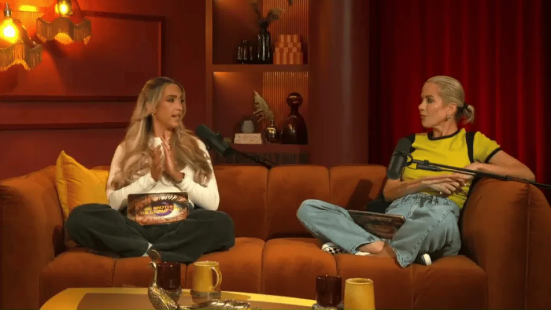 Two women seated on a rust-colored couch with microphones in front of them.