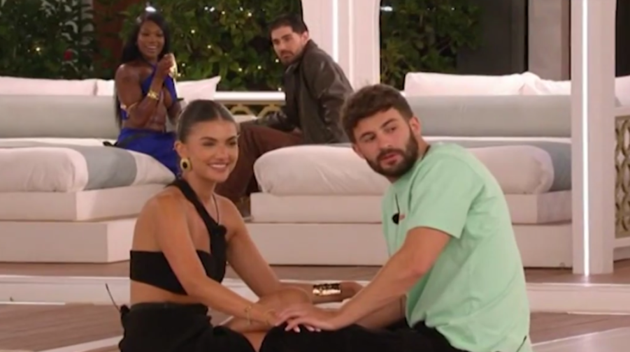 Two people sit on the floor holding hands while two others watch from couches behind them.