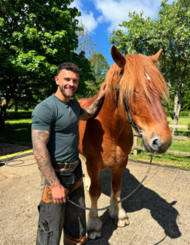 A smiling man, identified as a farrier by his attire, stands next to a horse with a long mane.