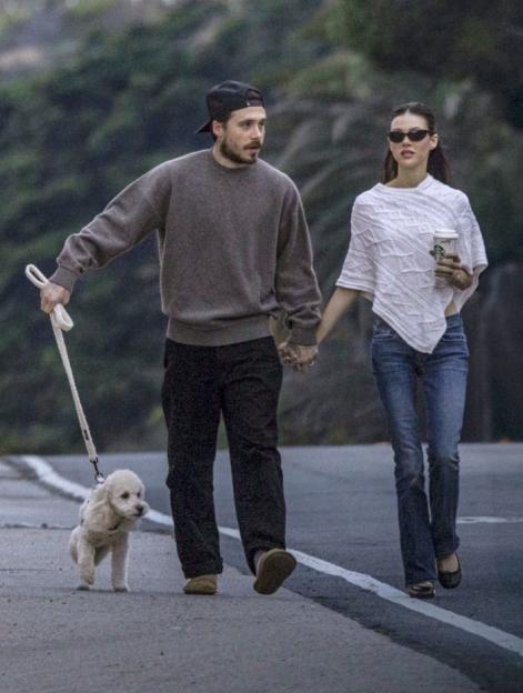Brooklyn Beckham and Nicola Peltz Beckham holding hands while walking a small white dog.