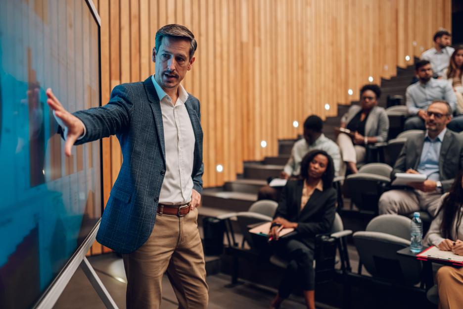 A speaker presenting to a diverse audience in a conference hall, gesturing at a large screen displaying information.