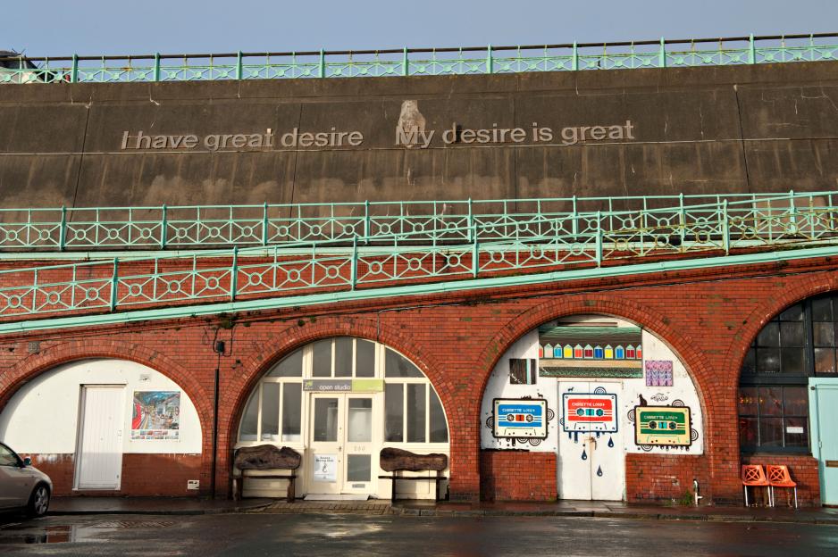 Retail outlets under brick arches on Madeira Drive in Brighton, UK, with a concrete wall above featuring the inscription "I have great desire My desire is great."