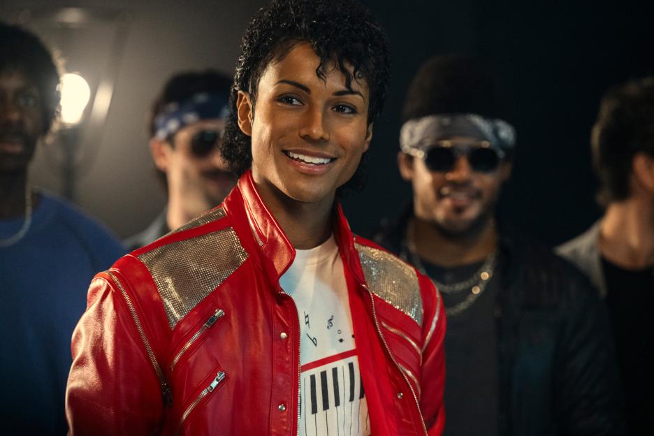 Jaafar Jackson as Michael Jackson in a red jacket with silver accents, smiling with a piano key graphic on his shirt.